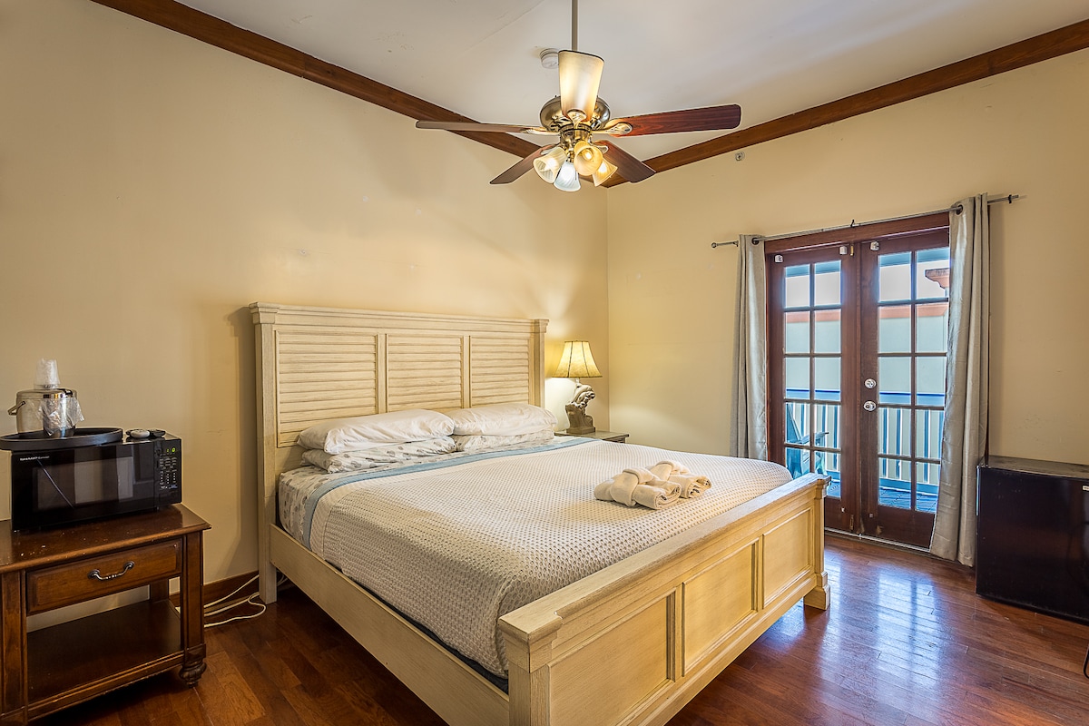 A spacious bedroom features a king-size bed with a light-colored headboard and soft linens. A ceiling fan adds to the comfort. There is a small dining area with a lamp, a microwave, and a mini fridge. Double doors open to a balcony with natural light flooding the room.