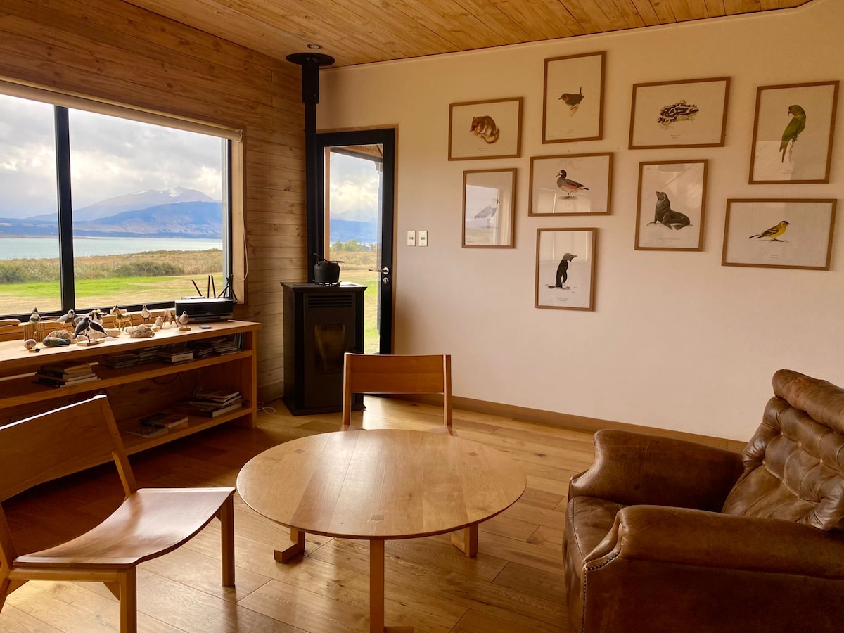 A cozy interior space features a light wood ceiling and walls. A large window offers views of the outdoors. A small wooden round table is surrounded by two simple chairs. A comfortable brown armchair is positioned nearby, and wall-mounted frames display various animal illustrations.