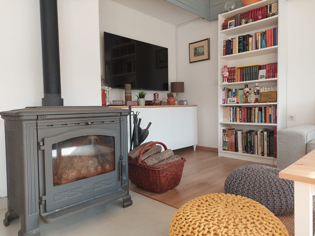 A cozy living area features a traditional wood stove, surrounded by a basket of logs for warmth. A neutral sofa is complemented by textured poufs, and a bookshelf filled with books is visible in the background. A modern television sits mounted on the wall.