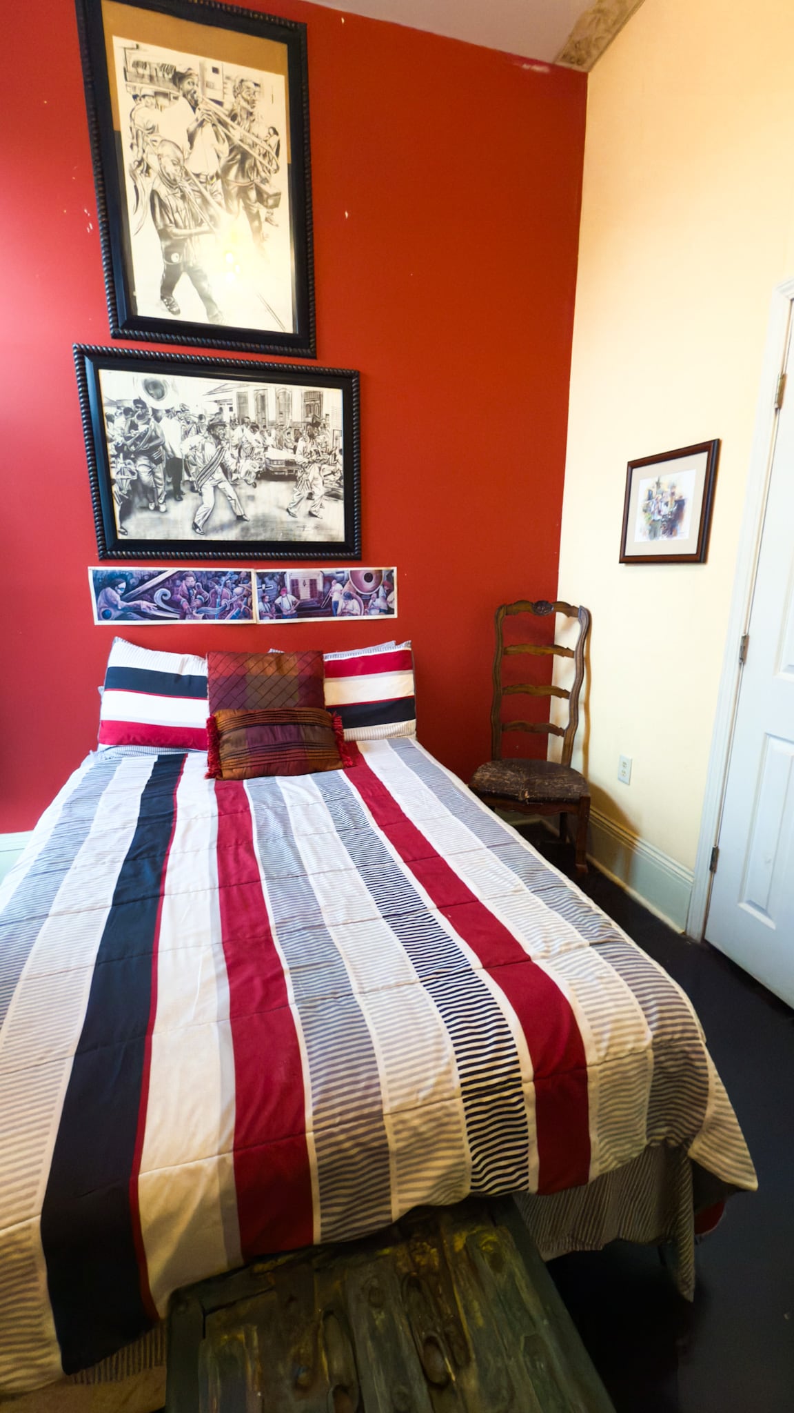 A cozy sleeping area is defined by a striped bedspread in red, white, and navy. Above the bed, framed black-and-white artwork adds character to the red accent wall. A wooden chair and side table complement the space, providing additional seating options.