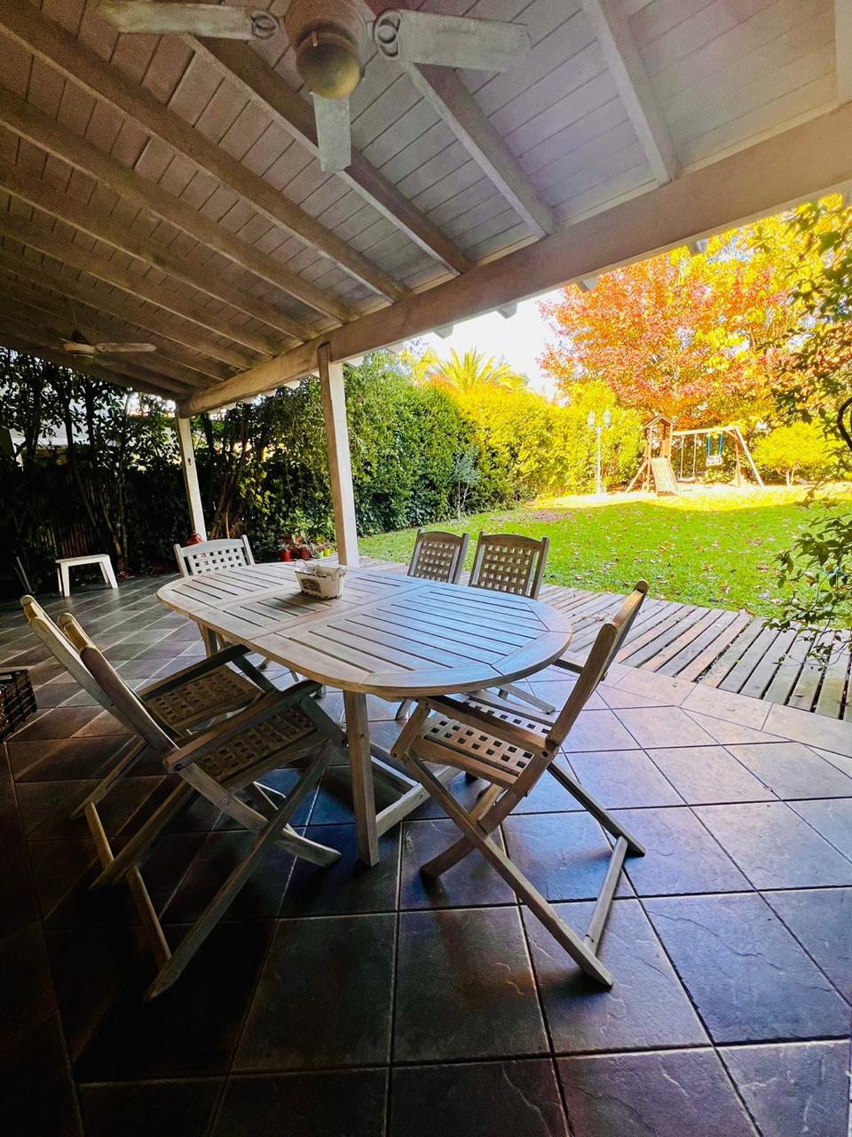 An outdoor dining area features a large table surrounded by six folding chairs, set under a covered patio. A view of a green garden is visible in the background, showcasing trees and a playset. Natural light filters through the space.