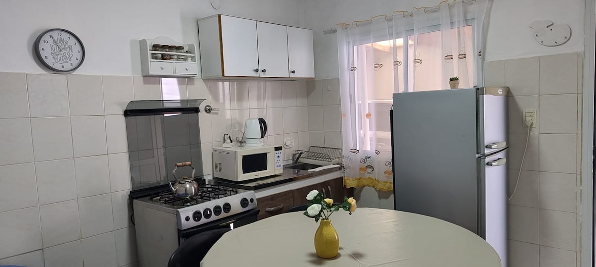 A functional kitchen space is equipped with essential appliances, including a stove, microwave, and refrigerator. A round dining table is centered, adorned with a small vase of flowers. Natural light enters through a window, and a clock is mounted on the wall.