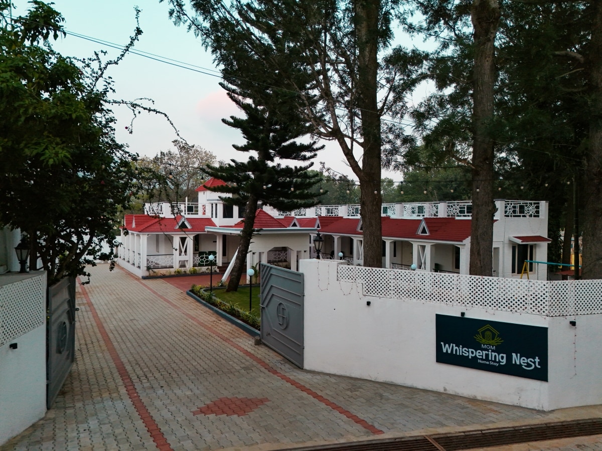 A charming villa complex is seen from the entrance, featuring a red-roofed building surrounded by tall trees. A paved pathway leads to the main entrance, bordered by well-maintained greenery. The sign for 'Whispering Nest' is displayed prominently, welcoming guests to the property.