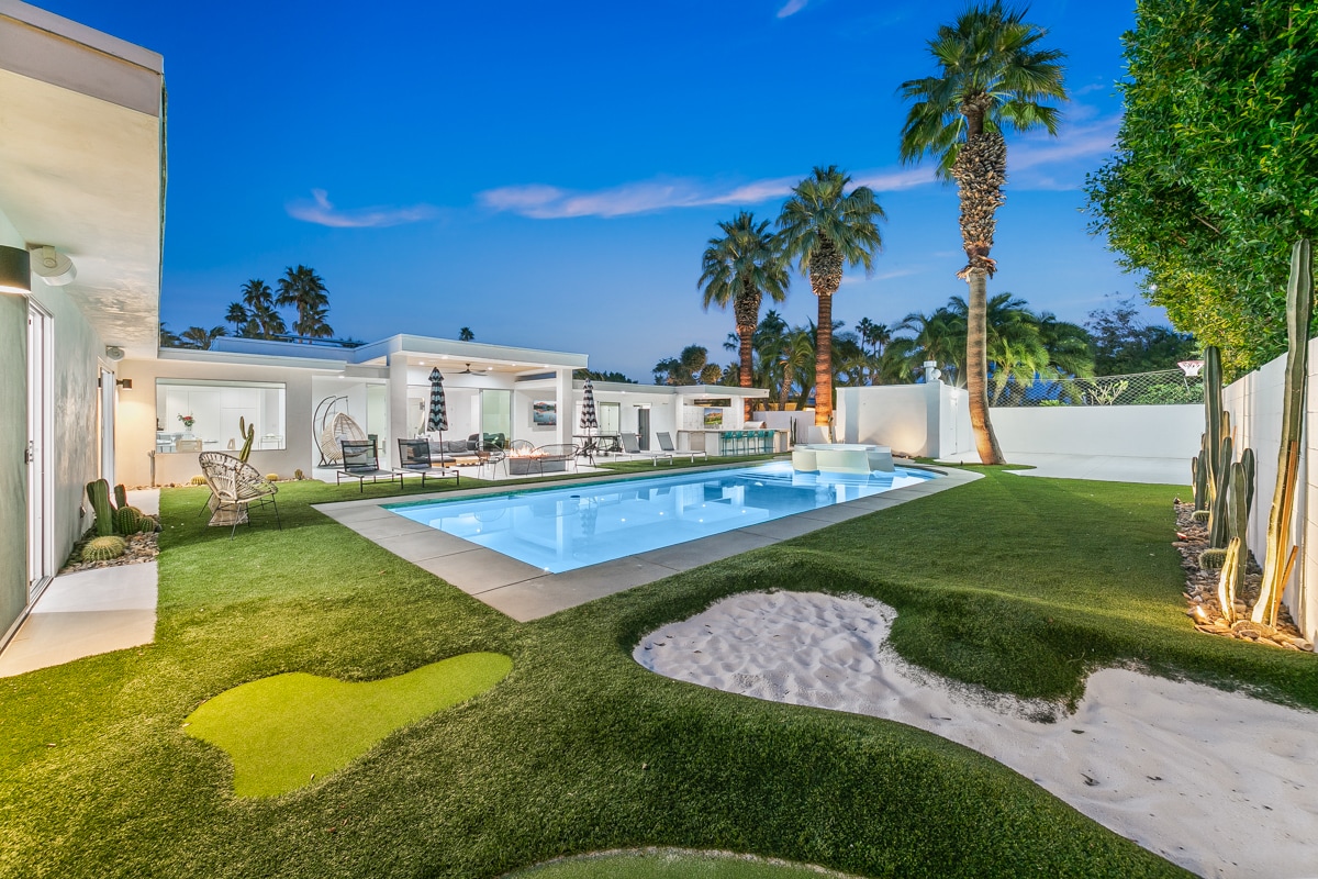 An expansive outdoor area features a modern swimming pool surrounded by lush green grass and palm trees. Comfortable seating arrangements are visible along the sides, complemented by organized landscaping elements, including low-maintenance sand areas and decorative plants. The backdrop showcases a clear evening sky.