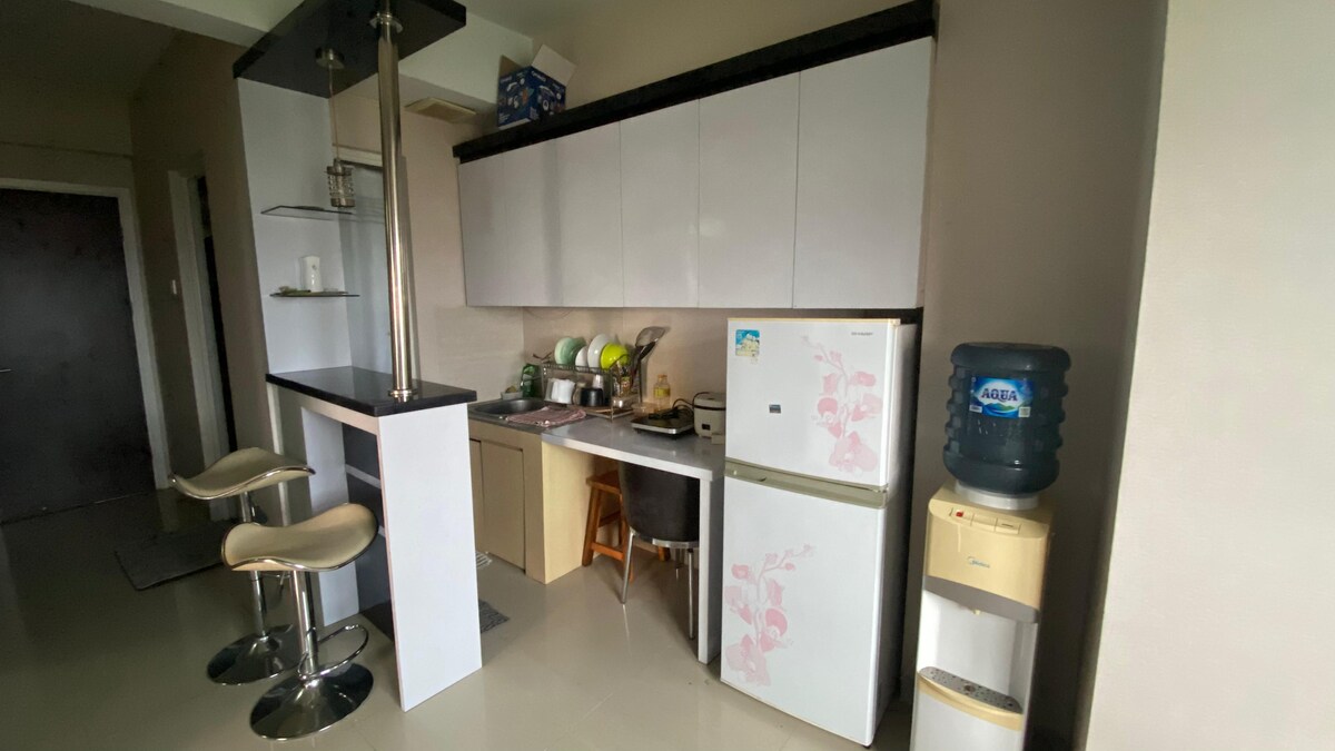 A compact kitchen area is visible, featuring white cabinetry and a small bar with two stools. A refrigerator stands beside a water dispenser, while an array of kitchen appliances is arranged on the countertop. Natural light fills the space.