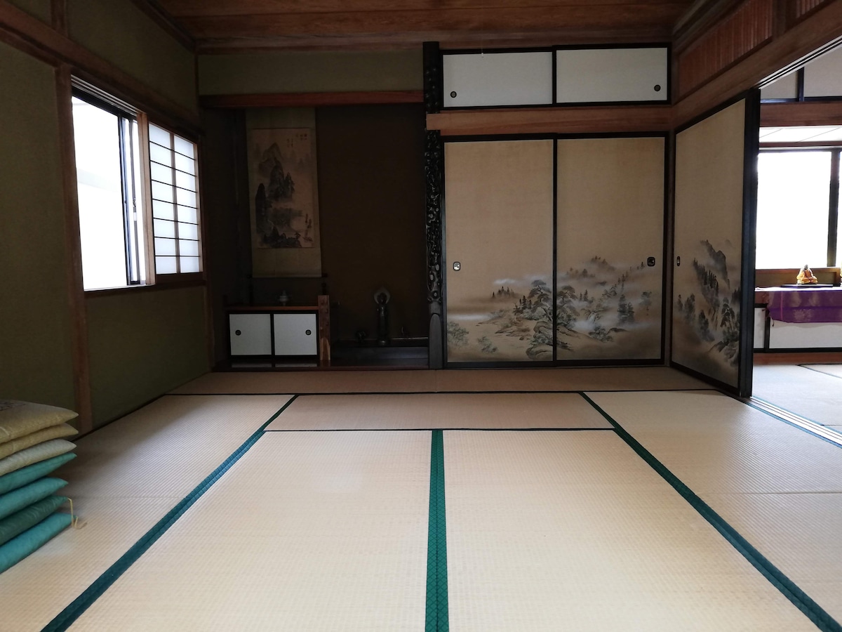 The traditional room features tatami mat flooring and sliding shoji doors adorned with muted artwork. One corner holds a decorative alcove, while a small storage compartment is integrated into the wall. Natural light enters through the windows, illuminating the serene space.