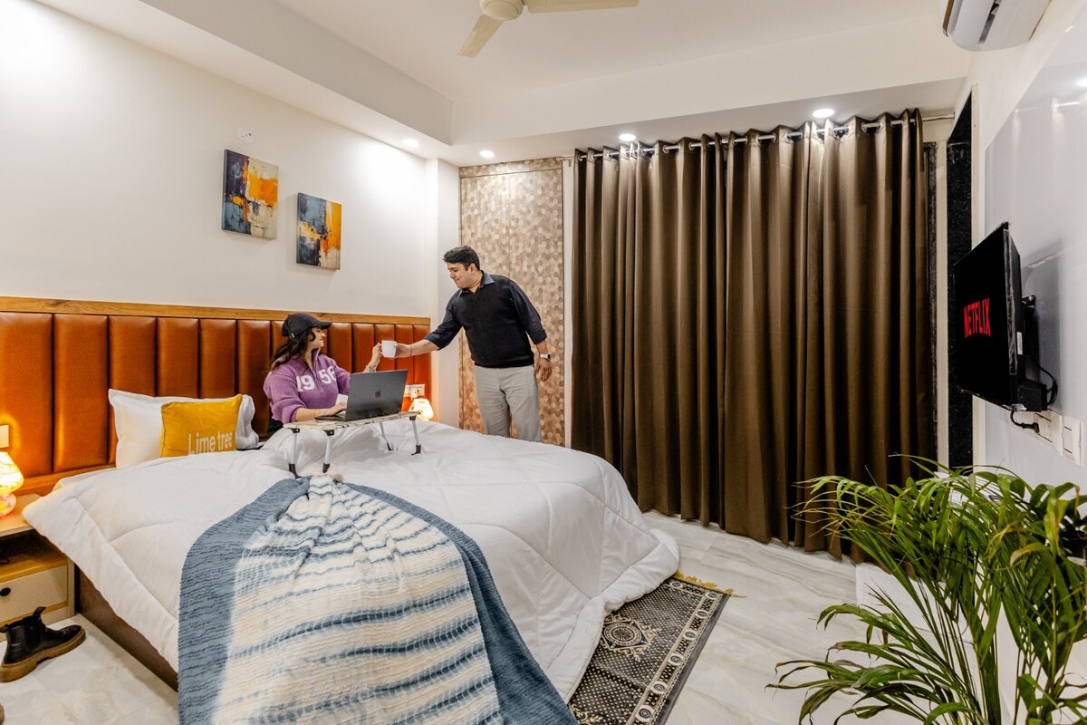 A well-lit bedroom features a spacious bed layered with a soft blanket and decorative pillows. A person is seated at a laptop, while another individual stands nearby. Dark curtains frame the window and artwork adds a touch of colour to the room's walls.