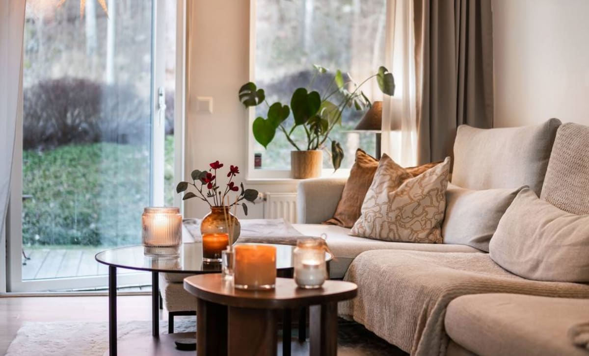 A cozy living area features a light-colored sectional sofa adorned with decorative pillows. Two wooden coffee tables display candles, providing soft lighting. Large glass doors lead to a green outdoor space, enhancing the room's natural light and connection to the outside.