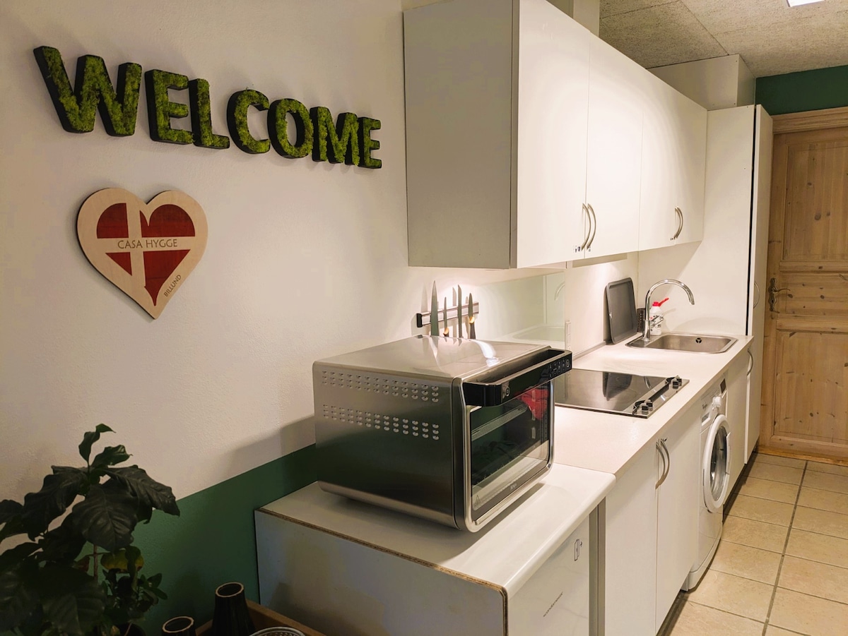 A clean and functional kitchen area is shown, featuring white cabinetry and countertop. A microwave, stove, and sink are visible. A washing machine is integrated into the cabinetry. A green 'WELCOME' sign and heart-shaped decor add a warm touch to the space.