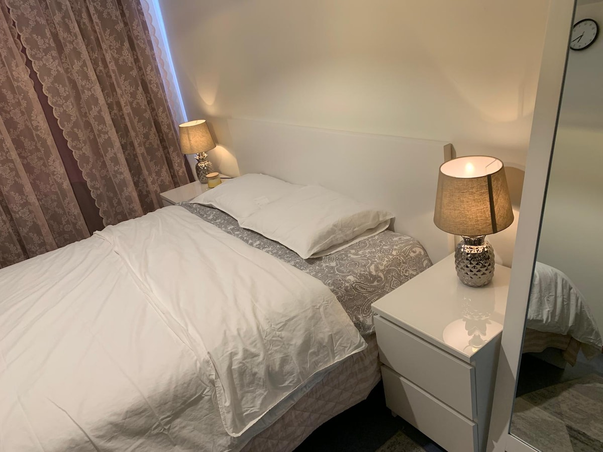 A cozy bedroom features a spacious bed with neutral-toned bedding, complemented by a decorative grey throw. A bedside table with a lamp provides soft lighting, and a large mirror reflects the warm ambiance. Curtains frame a window that allows natural light to enter.