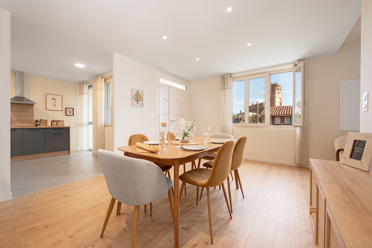 A modern dining area features a round wooden table set for a meal, accompanied by four stylish chairs. Natural light enters through large windows, illuminating the light-toned wooden floor and walls. The adjacent kitchen is visible, showcasing sleek cabinets and a stainless-steel range hood.