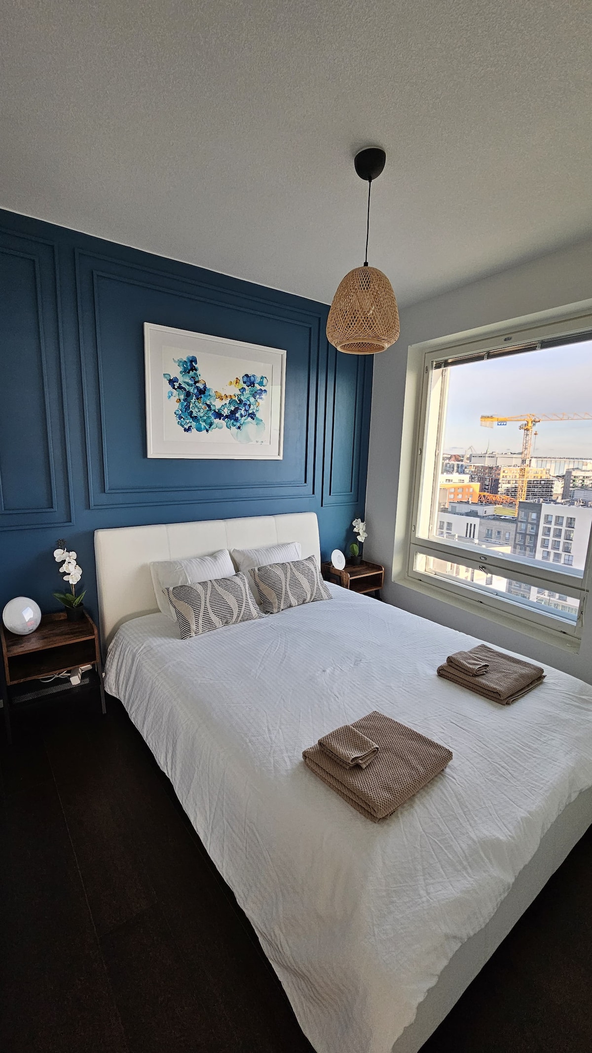 A serene bedroom features a spacious bed with light bedding, flanked by two nightstands with lamps. A blue accent wall adds depth, while a colorful piece of art adorns the wall above the bed. Large windows provide expansive views of the city below.