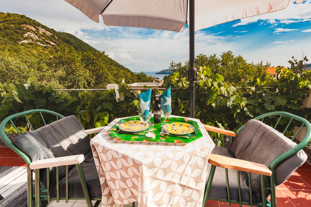 A private terrace features a round table set for two under a large umbrella. Two green chairs with grey cushions are positioned around the table, which is adorned with a floral tablecloth, plates, and glasses. A lush green backdrop of hills and distant views of the sea are visible.