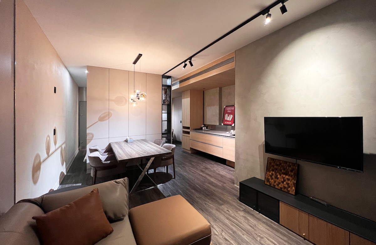 An open living space features a modern design with a cozy sectional sofa and a wooden dining table. The kitchen area is equipped with contemporary cabinetry. A flat-screen television is mounted on the wall, while soft lighting creates an inviting atmosphere.