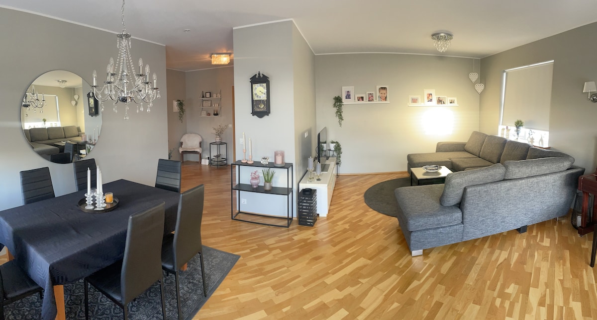 A spacious living and dining area features a black dining table with four chairs and a decorative centerpiece. A large gray sectional sofa is positioned near a soft rug, complemented by natural light from a window. A decorative mirror and wall art adorn the walls.