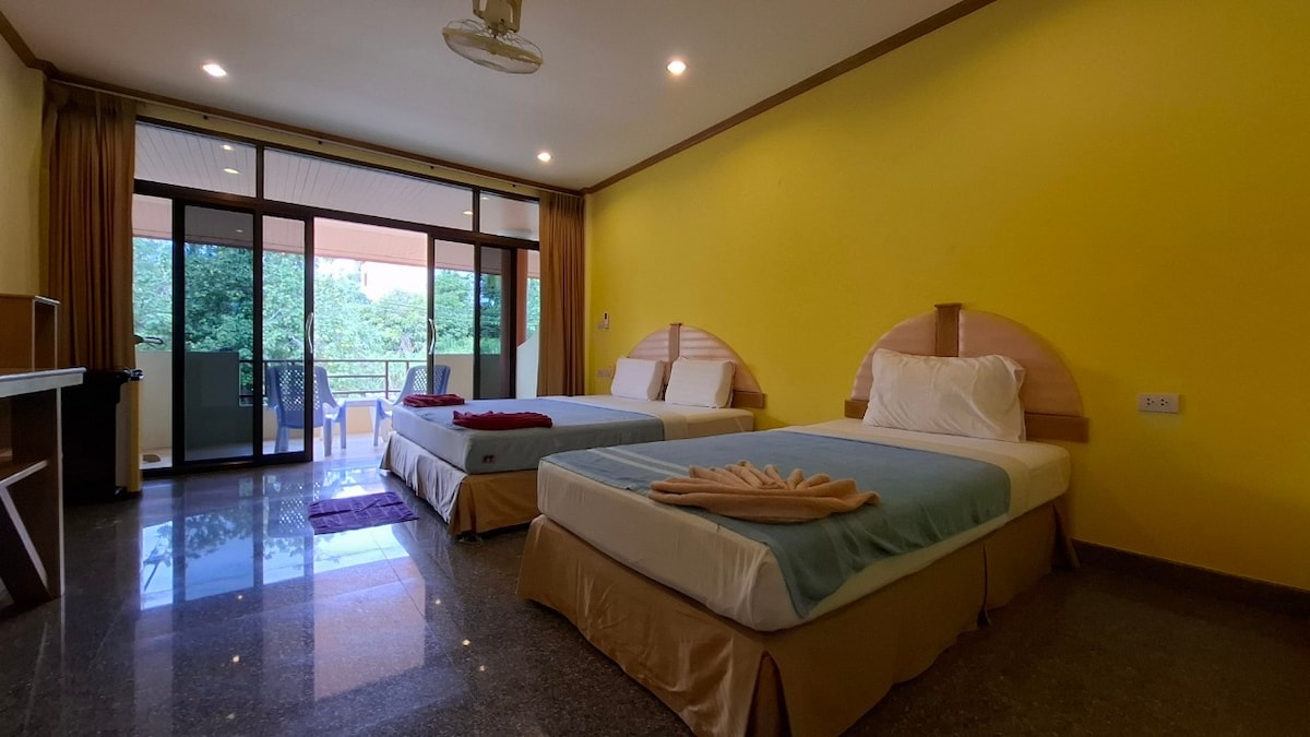 The spacious room features two inviting beds, each with soft linens. Large glass doors lead to a balcony, allowing natural light to fill the space. Neutral decor is complemented by a bright yellow wall, creating a cheerful ambiance.