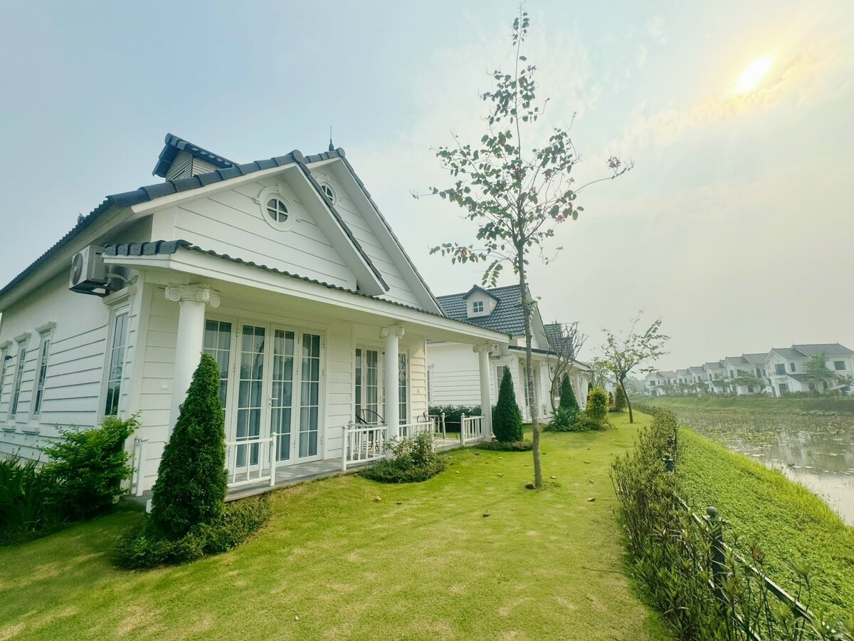 The exterior of a white, single-story villa is shown, featuring a row of large windows and a neighboring garden. Neatly trimmed hedges and young trees are visible on a grassy lawn, while a serene landscape extends toward a distant grouping of buildings.