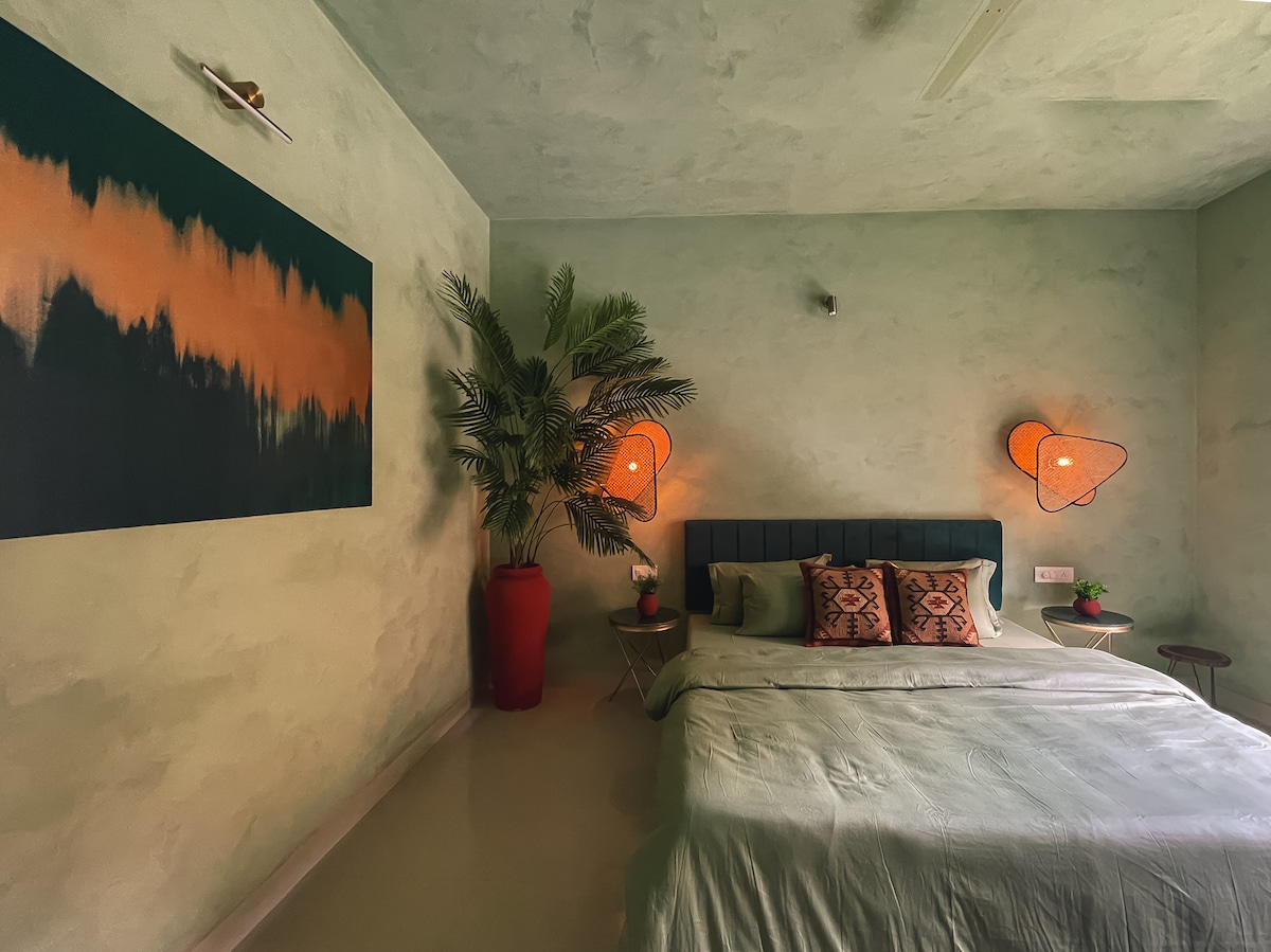The bedroom features a spacious bed with a muted green duvet and decorative pillows. A large painting with earthy tones adorns one wall, while potted plants add greenery. Two warm light fixtures softly illuminate the space, enhancing the calming atmosphere.