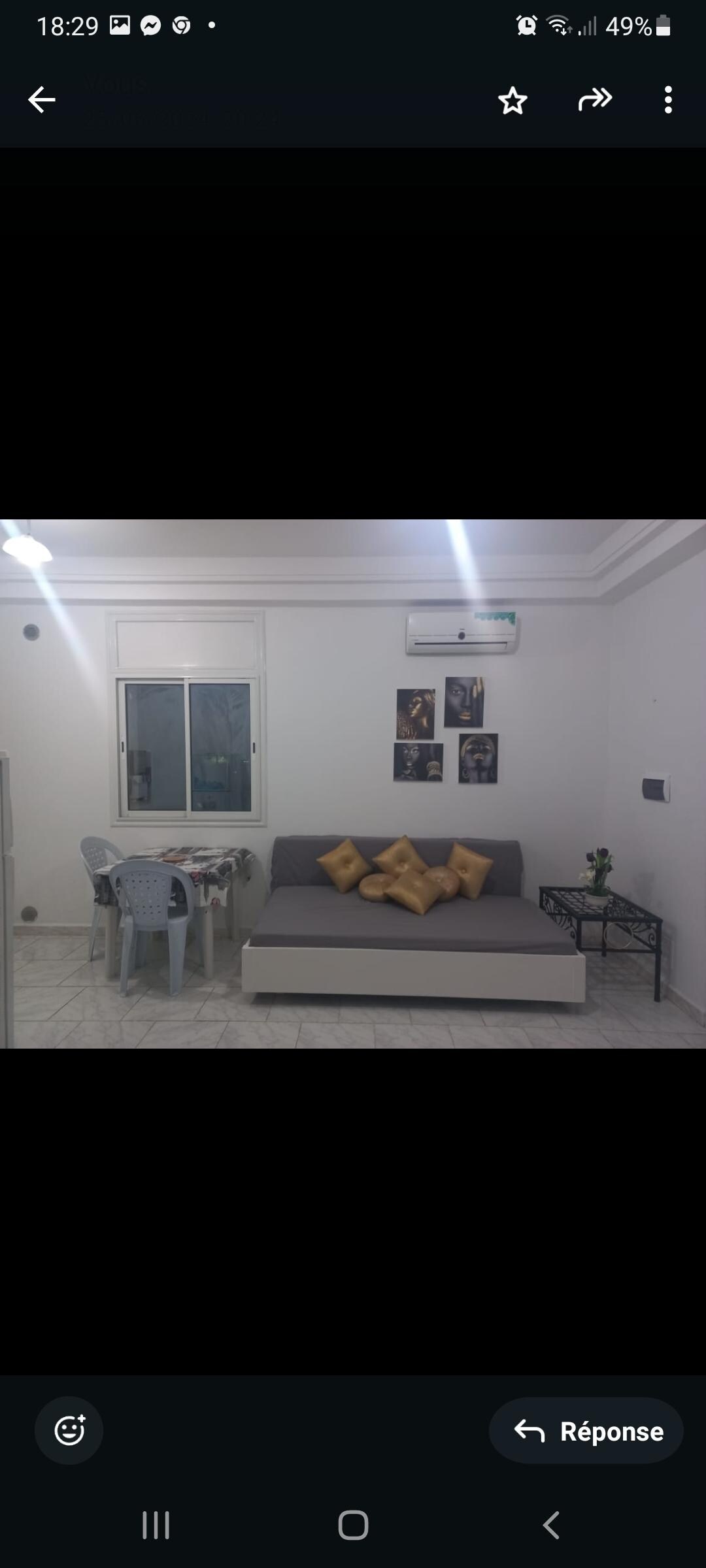 A well-lit living area features a gray sofa adorned with yellow cushions. A small dining table sits nearby, accompanied by four light blue chairs. On the wall, framed artwork adds character to the space, while a window allows natural light to brighten the room.