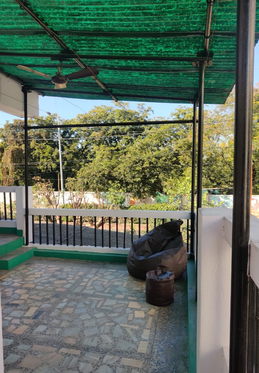 A shaded terrace area features a green canopy overhead, providing protection from sunlight. The floor is made of patterned stone, and a large bean bag is positioned in one corner. Surrounding greenery is visible, contributing to the peaceful outdoor setting.