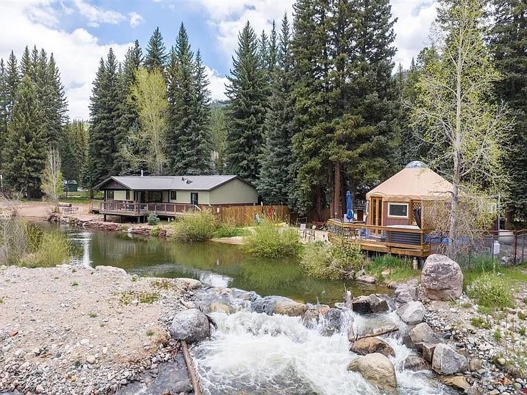 A rustic cabin is set beside a gently flowing brook, surrounded by tall pine trees. The structure features a welcoming deck area and a separate yurt nearby, both positioned to enjoy the serene natural landscape. Rocks border the water, enhancing the peaceful surroundings.