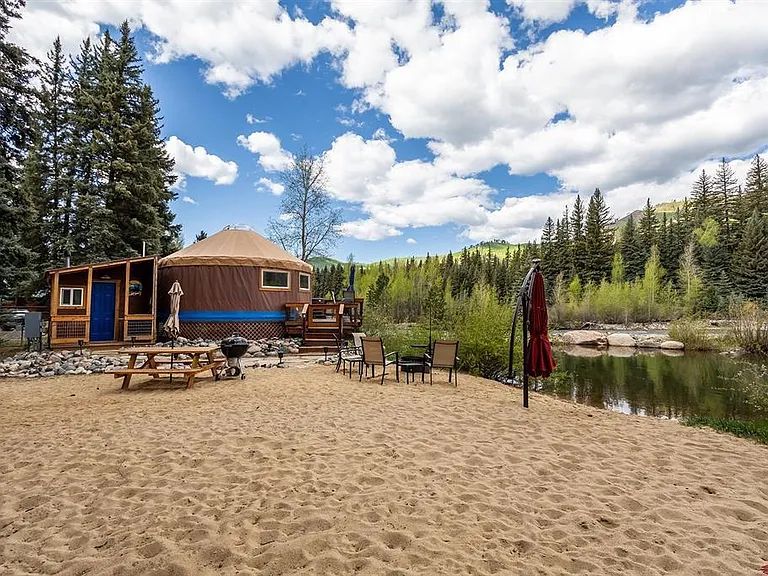 A rustic yurt is positioned near a sandy area, surrounded by trees and a calm body of water. Outdoor seating is arranged on the sand, featuring a picnic table and several chairs. Scenic mountains and a partly cloudy sky provide a serene backdrop.