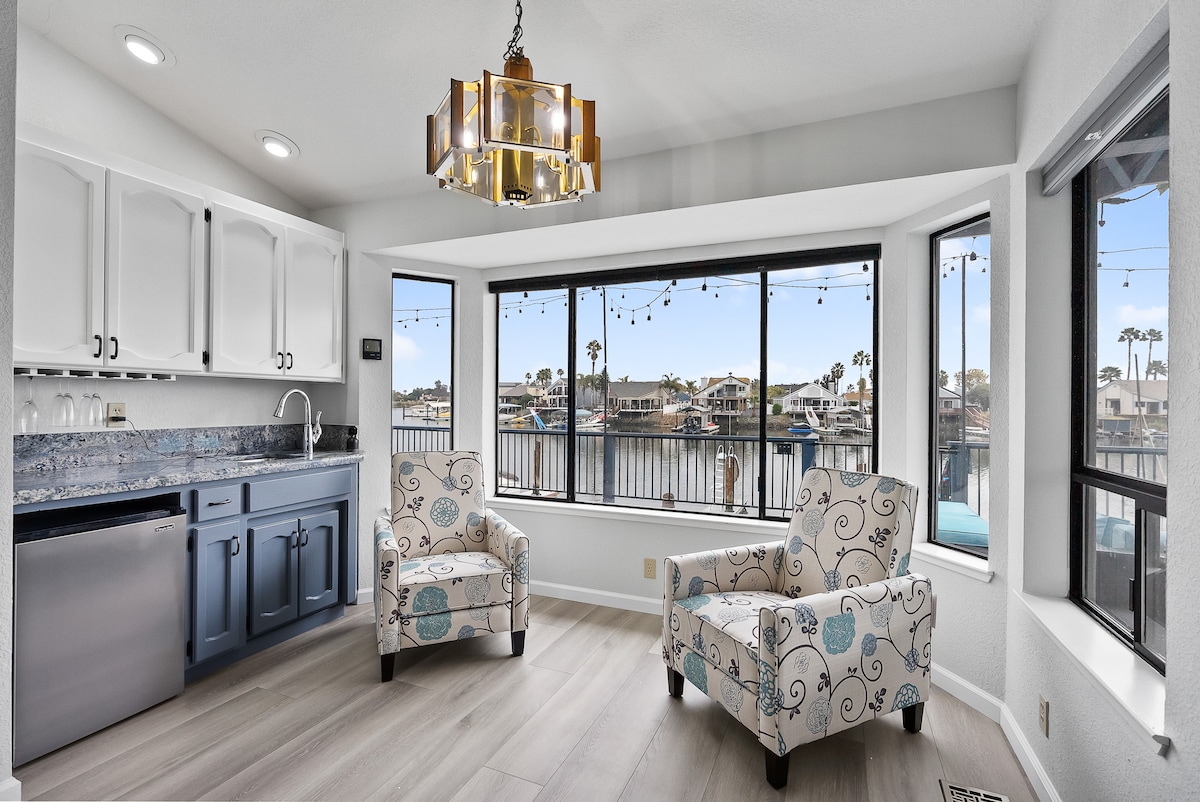 A bright corner nook offers two upholstered chairs with floral patterns, providing a cozy seating area. Large windows frame scenic waterfront views and allow ample natural light to fill the space. A modern kitchenette with white cabinetry and blue accents is also visible.