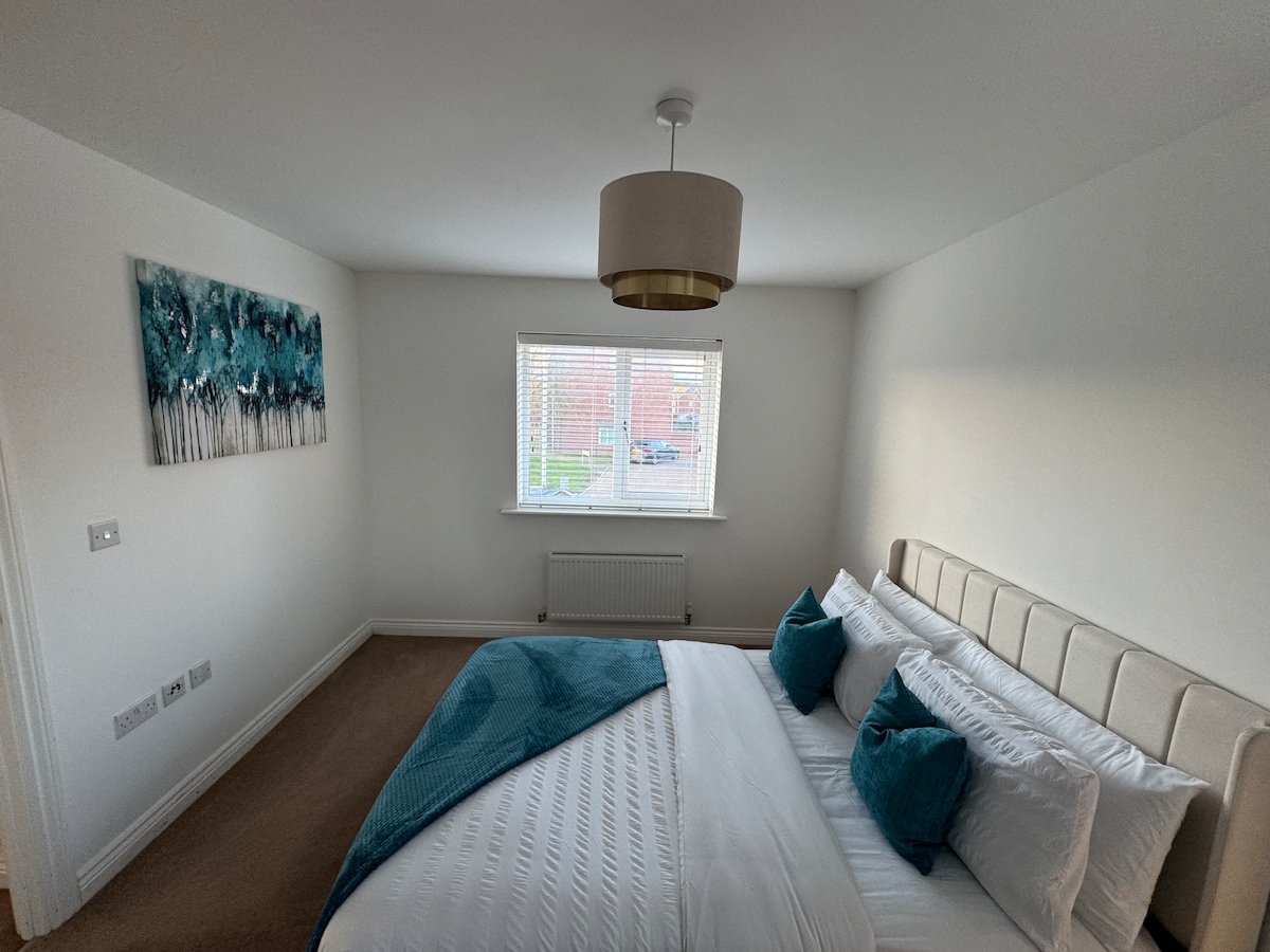 A well-lit bedroom features a spacious bed with a neutral-colored headboard, adorned with white linens and teal accent pillows. A piece of artwork depicting trees is displayed on the wall. Natural light enters through a window, enhancing the calming ambiance of the room.
