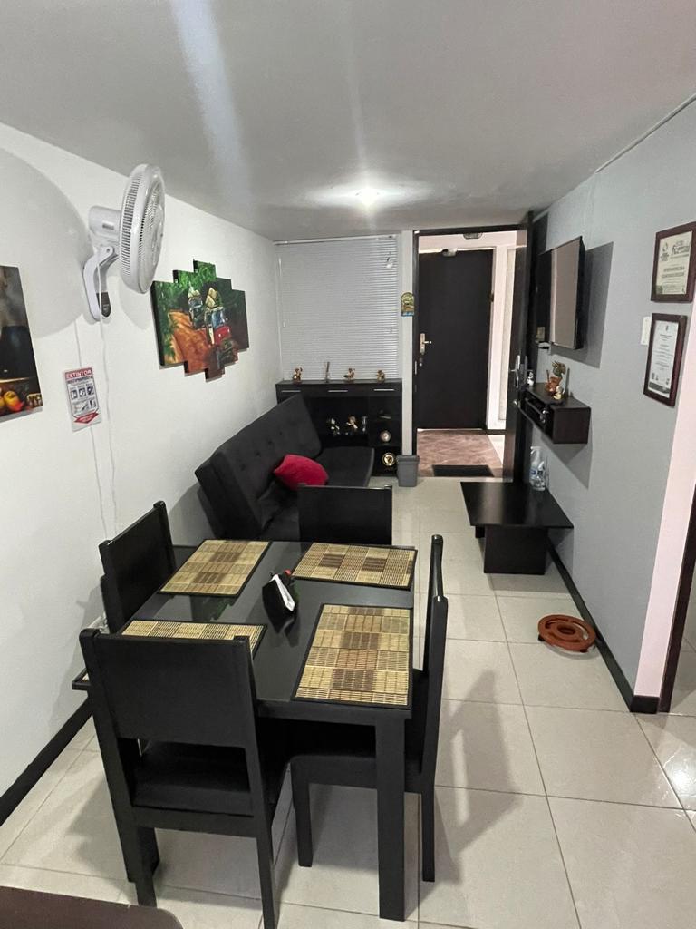 A modern living and dining area is presented, featuring a black upholstered couch and a square dining table set for four. The space is accented by framed artwork on the walls and a flat-screen TV mounted opposite the seating area. Light tiles enhance the room's brightness.