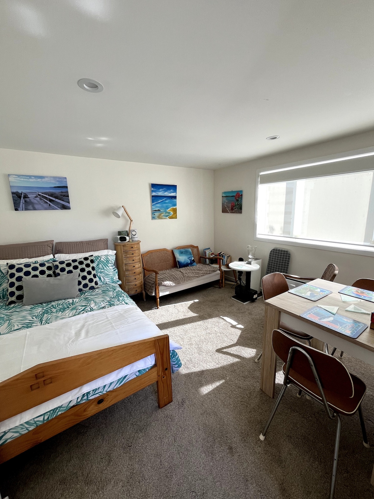 A well-lit room features a spacious bed adorned with patterned bedding and decorative pillows. A cozy seating area includes a small sofa and side table. A dining table with chairs is positioned nearby, and colorful artwork is displayed on the walls.