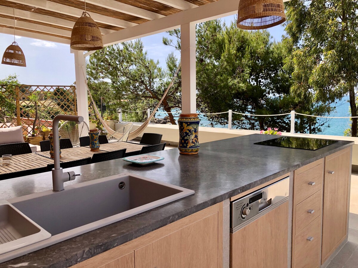 A spacious island kitchen features a stone countertop with a sink and dishwasher. Natural light filters through the overhead beams, illuminating the outdoor terrace with dining tables and comfortable seating. Lush greenery and glimpses of the sea are visible beyond the kitchen area.