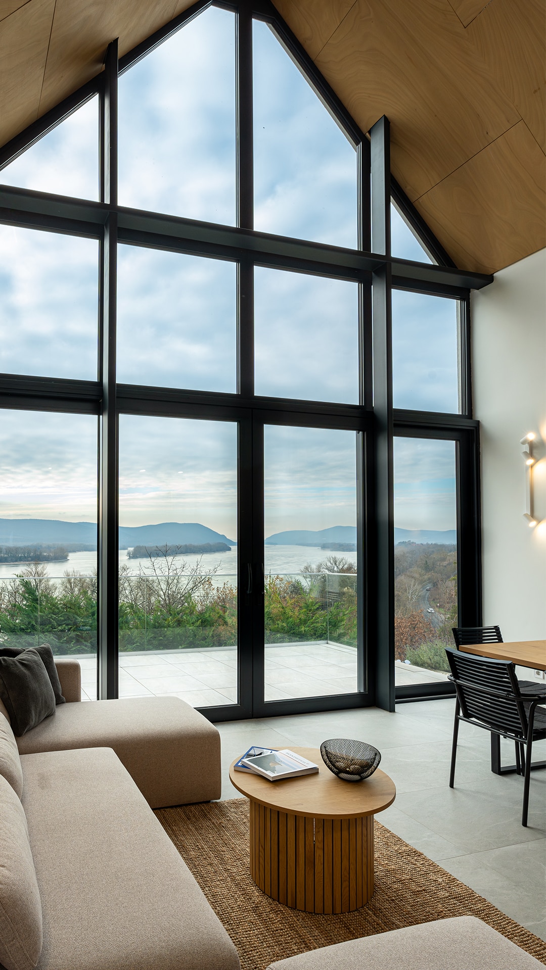 A modern living space is highlighted by large floor-to-ceiling windows, offering panoramic views of the landscape. A comfortable sofa sits adjacent to a wooden coffee table, while a dining area with black chairs is positioned nearby, allowing for both relaxation and dining.