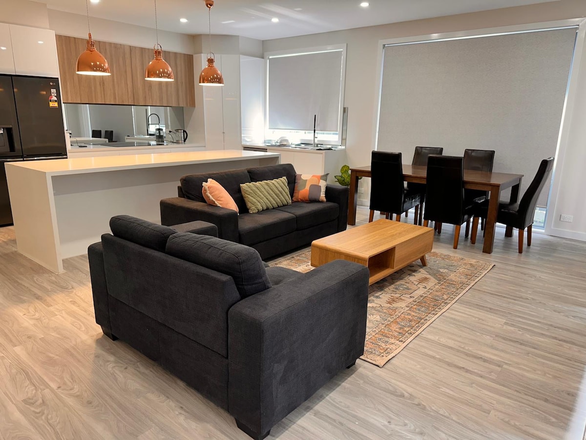 An open-plan living area is showcased, featuring a dark sofa with decorative cushions. A wooden coffee table rests on a patterned rug, while a dining table with six chairs is positioned in the background. Contemporary pendant lights hang above the kitchen counter.