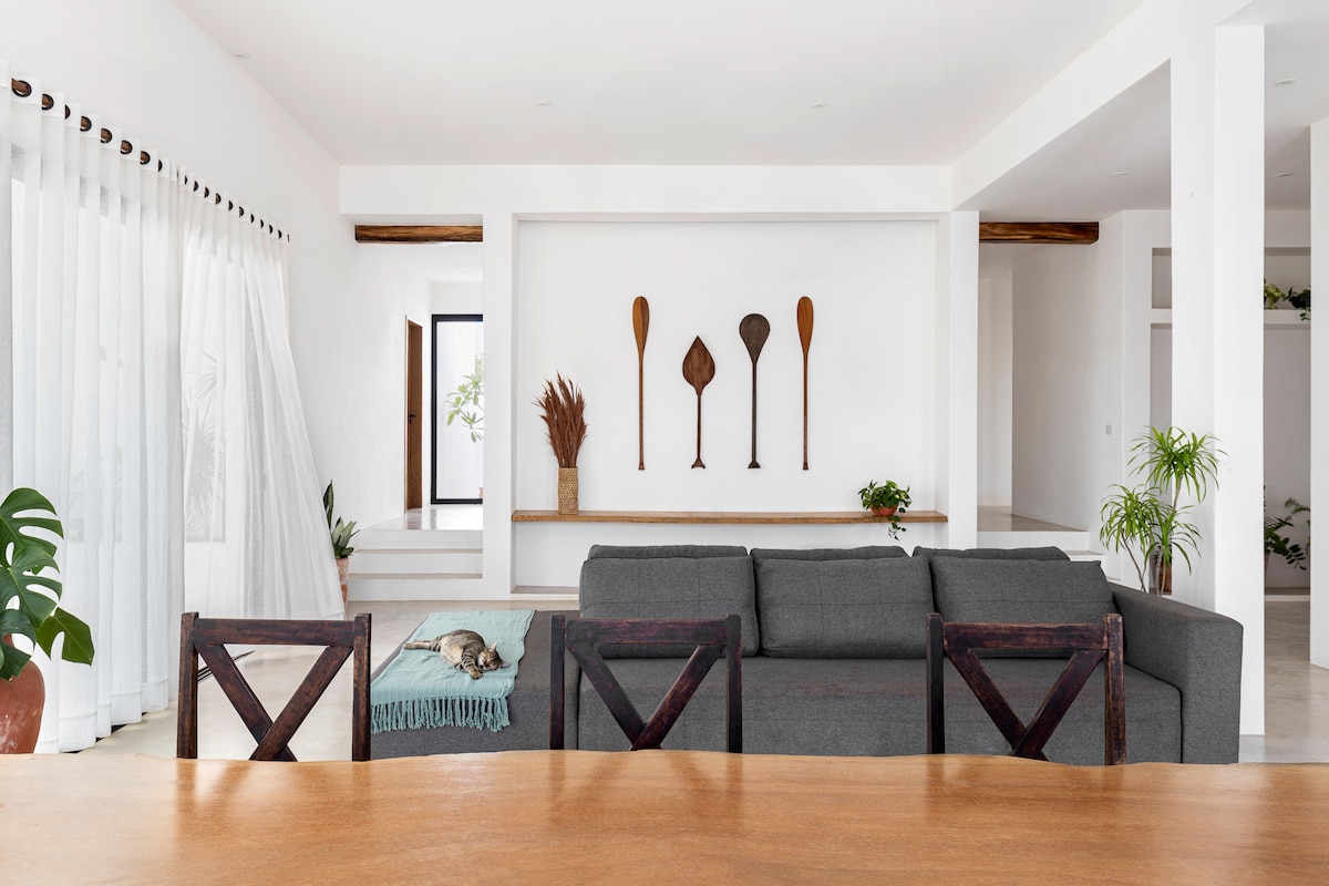 An open living area features a grey sectional sofa adorned with a blue throw, positioned towards a feature wall displaying decorative wooden paddles. Light-colored walls and a bright atmosphere enhance the spacious feel, while large windows invite natural light into the room.