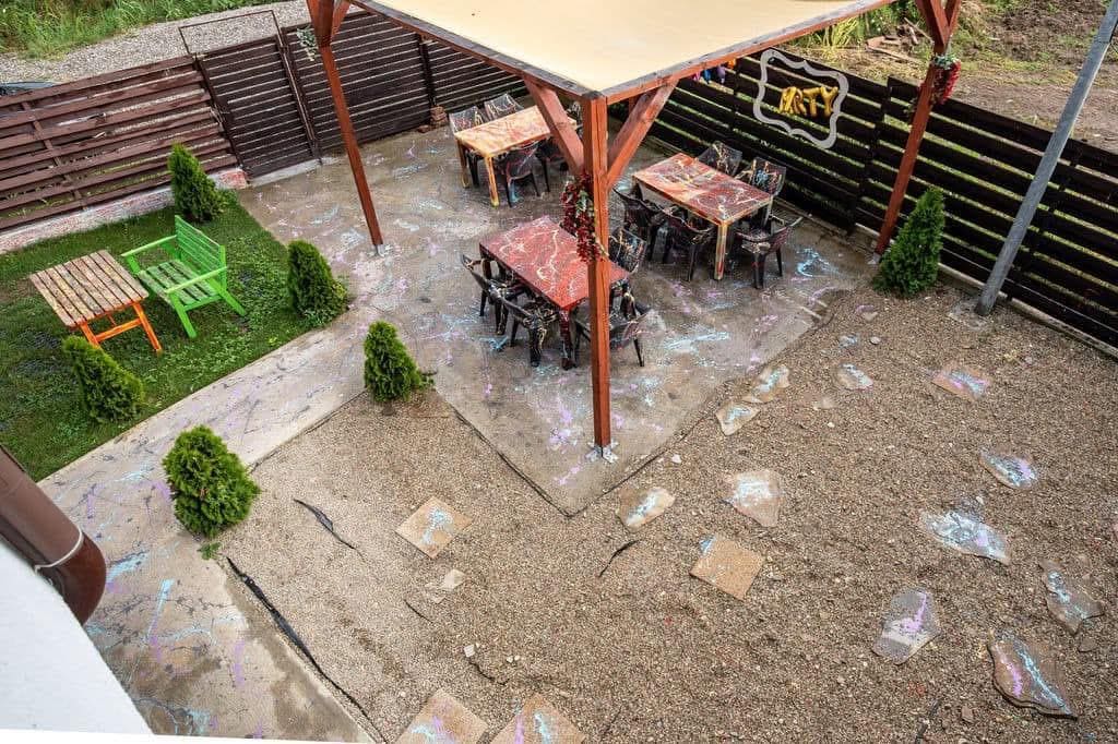 An outdoor area is showcased with multiple wooden tables and chairs arranged beneath a sheltered pergola. Greenery lines the space, complemented by two small shrubs. A pathway made of stones leads through a gravel surface, enhancing the inviting atmosphere of the gathering space.