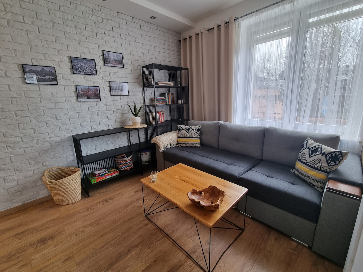 A comfortable living area features a grey sectional sofa adorned with patterned cushions, positioned near large windows that allow natural light. A wooden coffee table sits in front, accompanied by a decorative bowl. Bookshelves filled with various books and a plant add a welcoming touch.