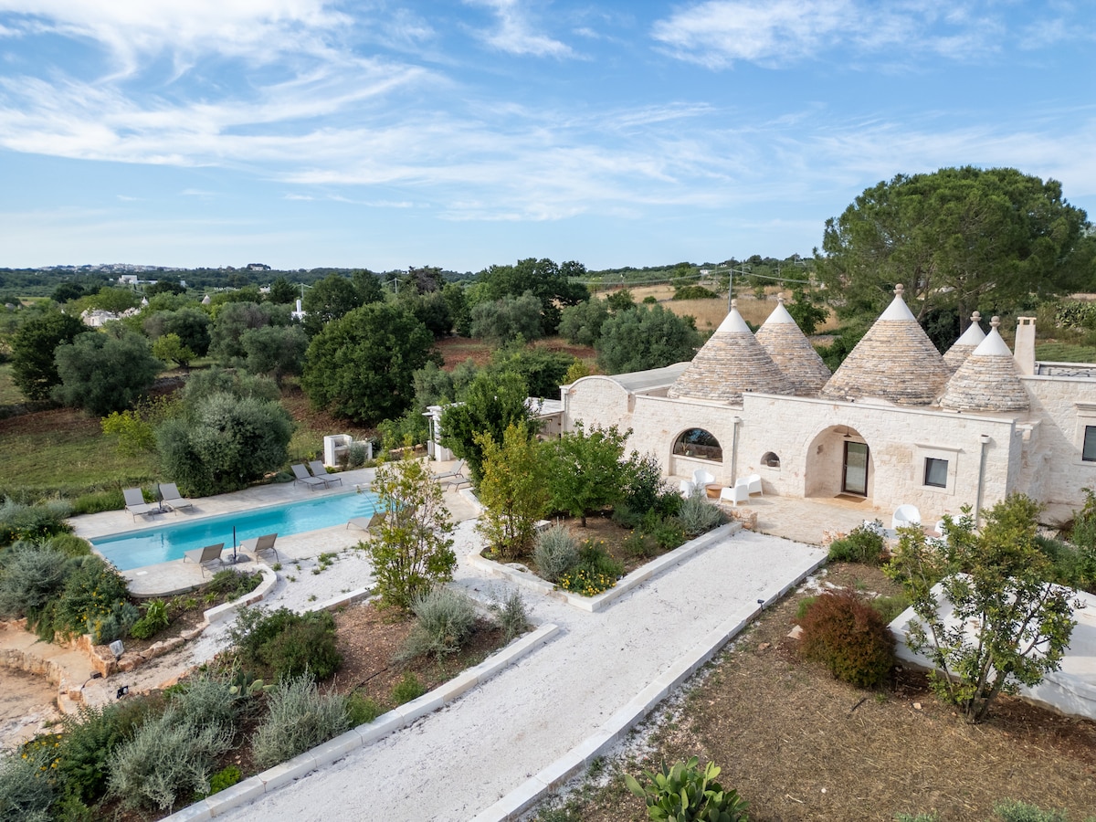 The view showcases a charming complex of trulli with conical roofs nestled among greenery and trees. A clear pool is visible beside the structure, surrounded by a well-kept garden. A white pathway leads from the entrance, enhancing the serene atmosphere of the countryside setting.