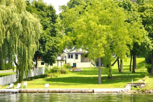 A spacious yard leads to the lake, bordered by lush trees and a white fence. The home is visible in the background, surrounded by greenery, with seating available for relaxation by the water's edge.
