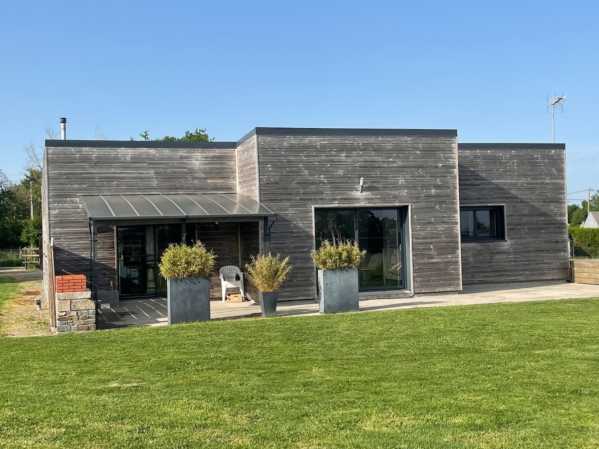 A modern one-story house is presented with a wooden exterior, complemented by large windows. A covered entrance with seating is visible, flanked by planters. The surrounding lawn provides a spacious outdoor area, enhancing the home's connection to nature.