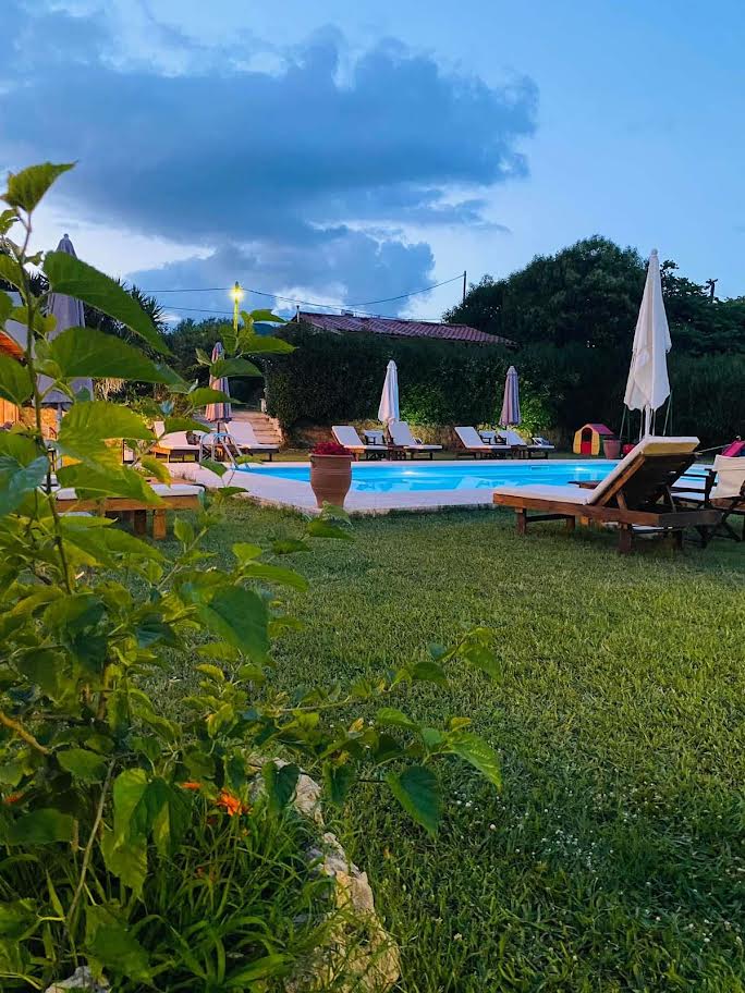 A tranquil outdoor space features a well-maintained grassy area surrounding a blue pool. Loungers are arranged around the pool, complemented by umbrellas. Lush greenery and flowering plants are visible in the foreground, contributing to a serene and inviting atmosphere during dusk.
