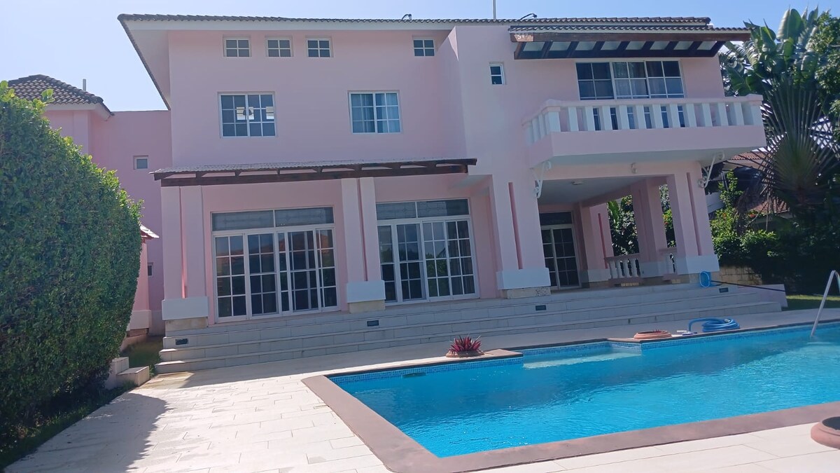 A spacious, two-story mansion is displayed, featuring a light pink exterior and multiple large glass doors. A tranquil swimming pool is set in the foreground, surrounded by stone tiles. Lush greenery lines the property, providing privacy and complementing the serene atmosphere.