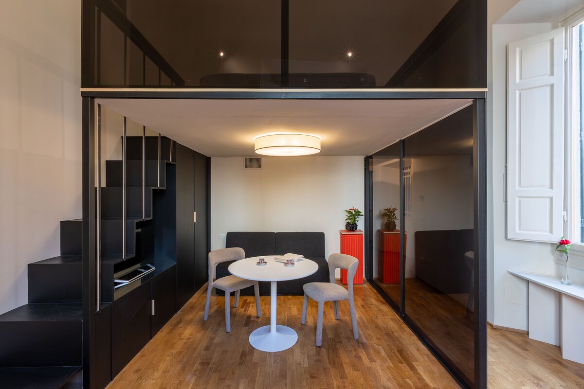 A modern living area features a circular white table surrounded by four light-colored chairs. A dark upholstered sofa is positioned against a glass partition. A statement light fixture hangs above, while natural light enters through large, partially open windows.