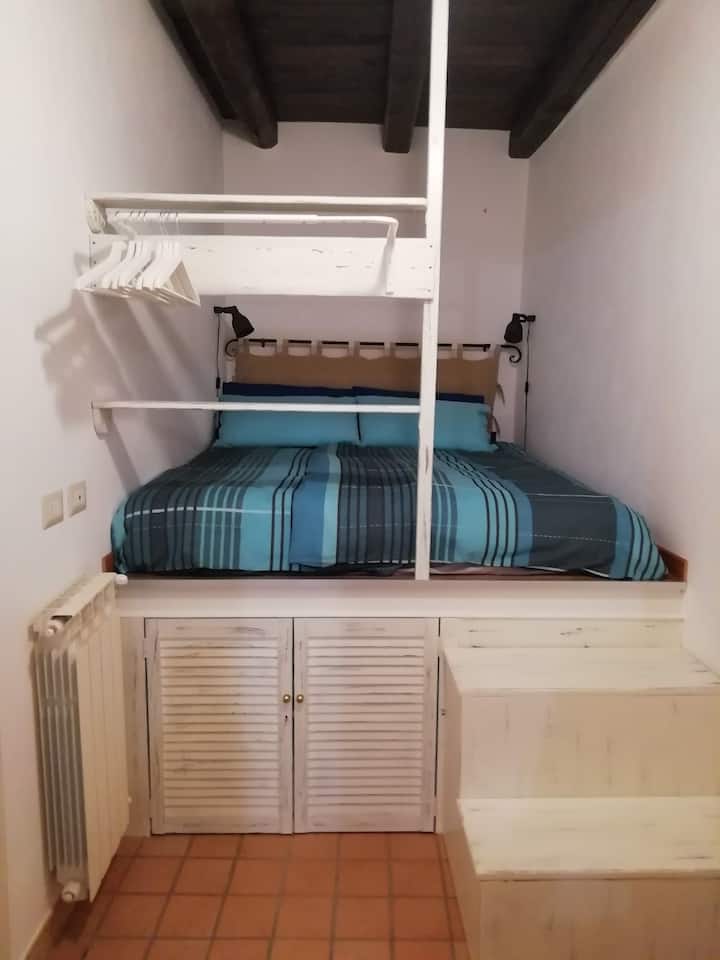  Აწეული საწოლი ორი საფეხურით, მატრასით პირდაპირ ხის იატაკზე, აღჭურვილია ქურთუკის საკიდით და ტელევიზორით, საწოლის თავსაფრის უკან ფანჯარა უზრუნველყოფს კარგ ვენტილაციას, ვენტილატორს კომფორტისა და სიახლის უზრუნველსაყოფად