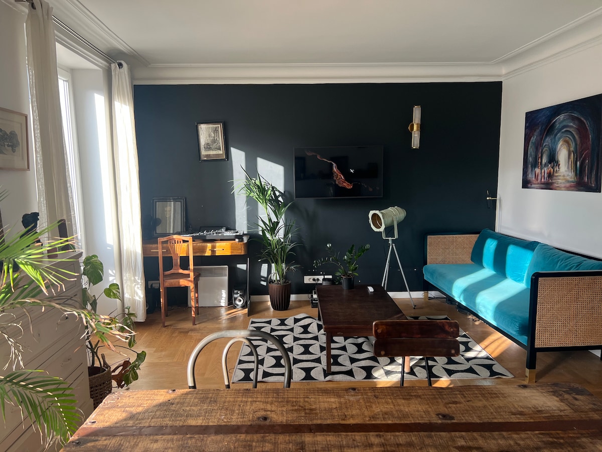 A living area is shown with a blue sofa and a wooden coffee table on a patterned rug. A television hangs on the wall opposite a desk with a chair. Natural light filters through the large windows, creating a bright atmosphere enhanced by green plants.