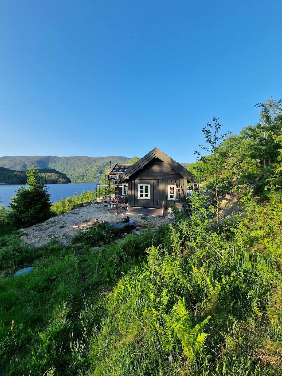 A newly constructed cabin is set against a scenic backdrop, positioned near a body of water. Lush greenery surrounds the cabin, with trees and shrubs visible. The structure features a sloped roof and multiple windows, while scaffolding indicates ongoing construction efforts.