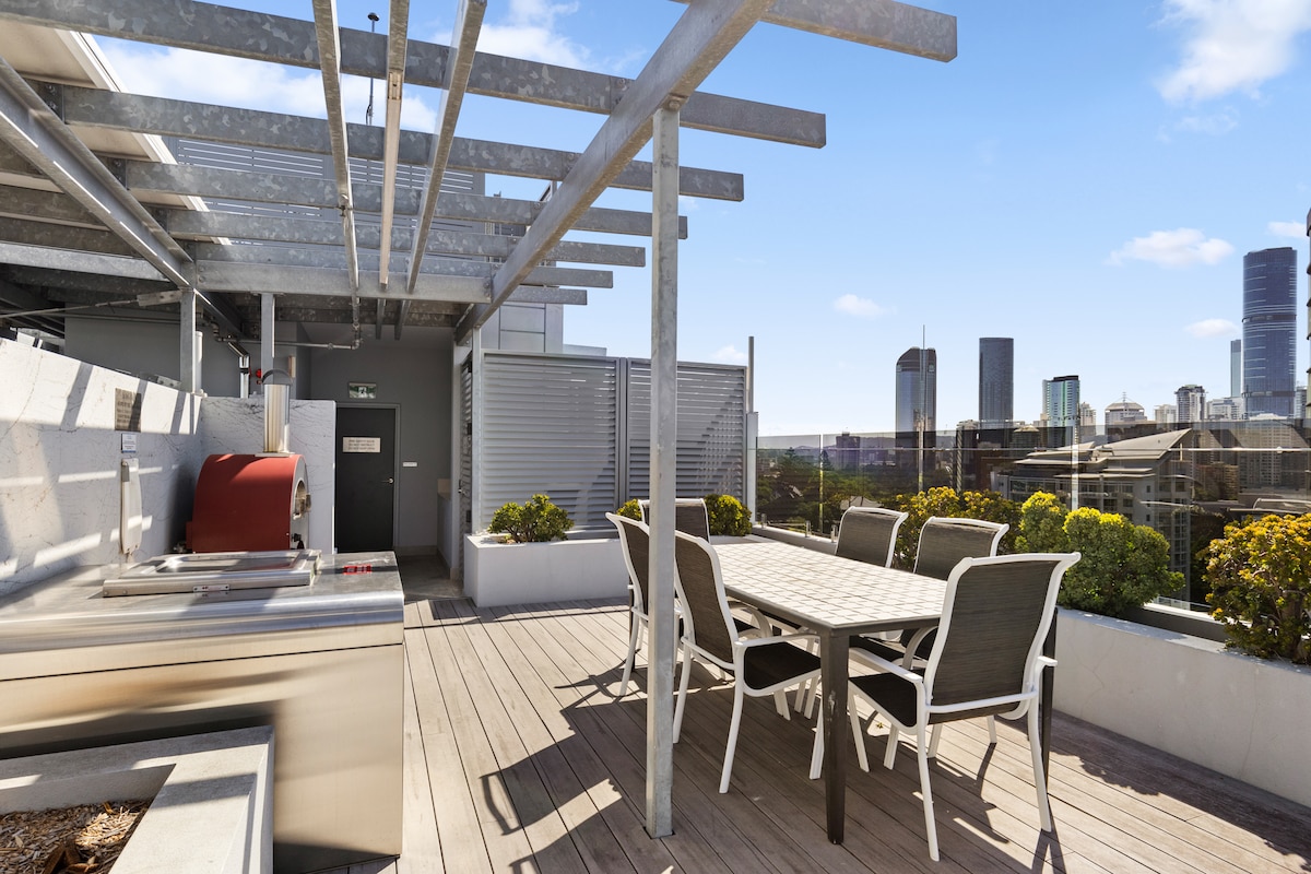 An outdoor dining area is offered on the rooftop, featuring a large table surrounded by several chairs. A grill is visible, situated among planters with greenery. The city skyline can be seen in the background, highlighting a blend of buildings against a clear blue sky.