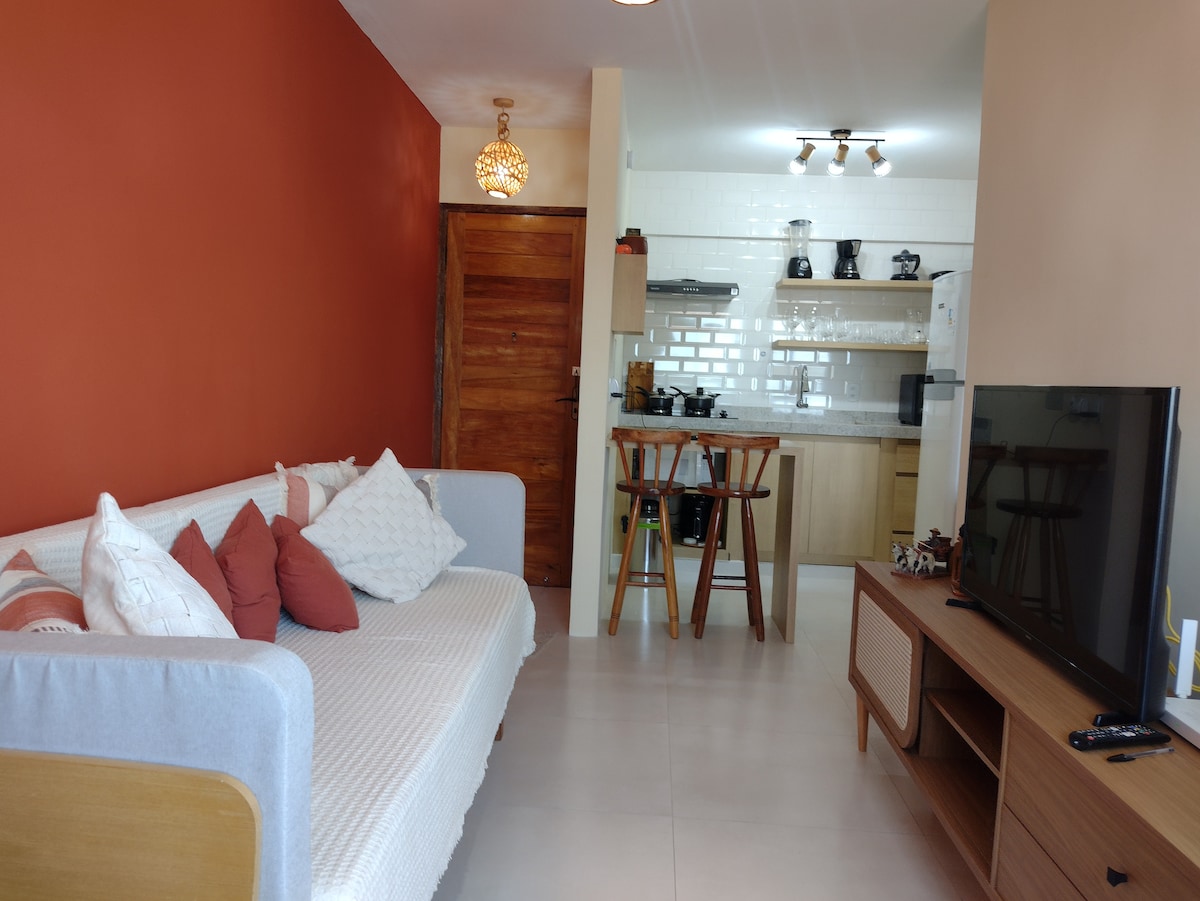A living area features a light sofa adorned with decorative pillows against an orange accent wall. A dining table with bar stools is positioned near an open kitchen, which is equipped with modern appliances. A flat-screen TV sits on a wooden console across from the seating area.