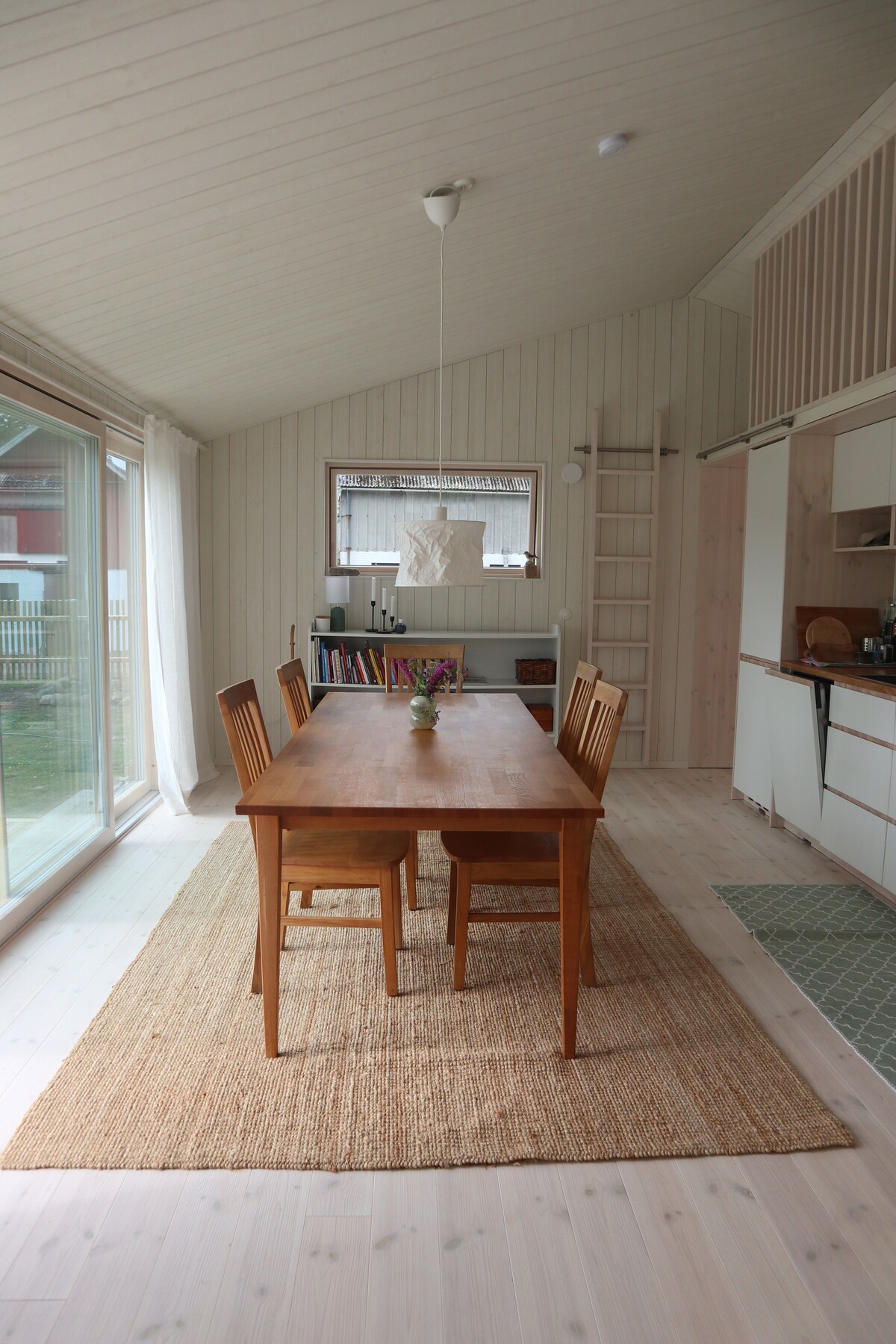 A spacious dining area features a wooden table surrounded by six chairs, illuminated by natural light from large sliding glass doors. A textured rug lies under the table, enhancing the neutral tones of the walls and ceilings. Kitchen elements are visible in the background.