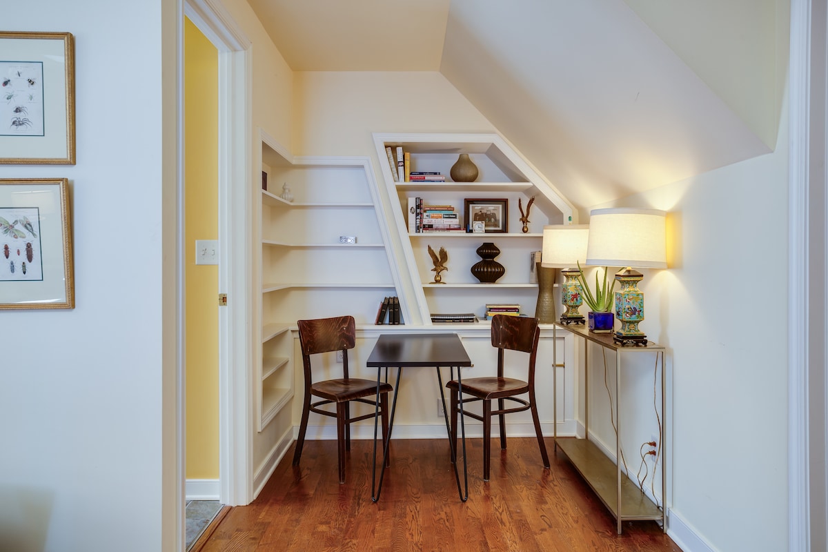 A dedicated workspace is featured in a well-lit corner, showcasing a small table paired with two wooden chairs. Shelves are lined with decorative items and books. A lamp provides additional illumination, and a small plant adds a touch of greenery.