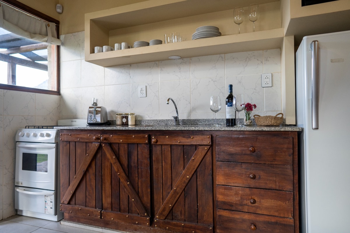 A kitchen area features wooden cabinetry with multiple drawers and an open shelf displaying dishware. Counter space is equipped with a wine bottle, drinking glasses, and a coffee maker. A compact stove and refrigerator are also present, offering essential appliances for meal preparation.